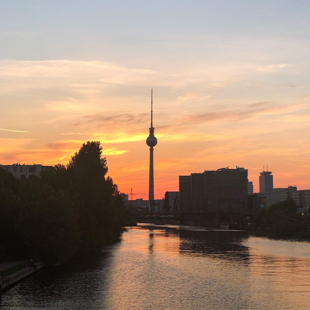 An orange sunset over a river behind the silhouette of Berlin's needle tower. 
