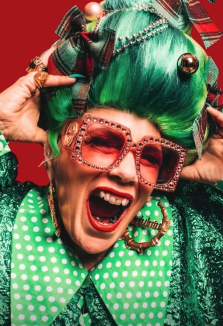 Woman with vibrant green hair, oversized orange glasses, and festive attire laughs energetically. The background is bold red, conveying a lively mood.