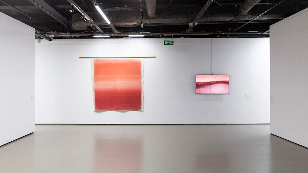 Abstract painting on a gallery wall with shades of red entitled Flow by Kim Taek Sang. Alongside the painting is a video screen showing the artists process to make the painting.