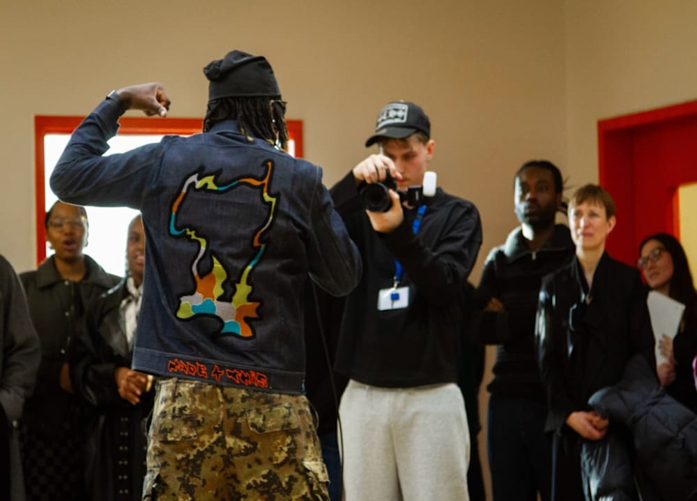 A photographer takes a shot of a person with their back to camera who is dressed in streetwear and appears to be performing while people look on.