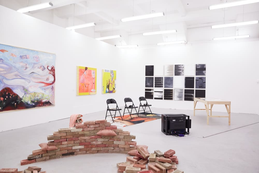 Room of art work by BA Fine Art Painting students. In the foreground is a low wall made of stacked, hand-made bricks. Behind this are three black fold-up chairs on a carpet facing a screen placed on the floor. To the right is a wooden tretle table. On the back wall is a colection of black, white and grey canvases hung together in a grid. On the left wall, two bright paintings in yellow and pink and a large canvas with a pale blue, black and red seascape.