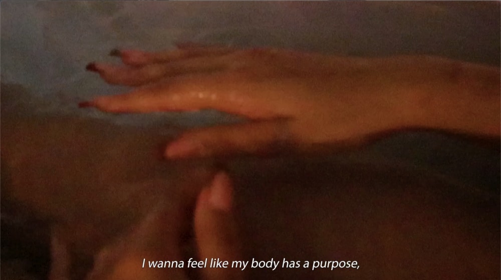 A still of a film showing a hand with the words 'I wanna feel like by body has purpose'. The person has acrylic nails with red tips.