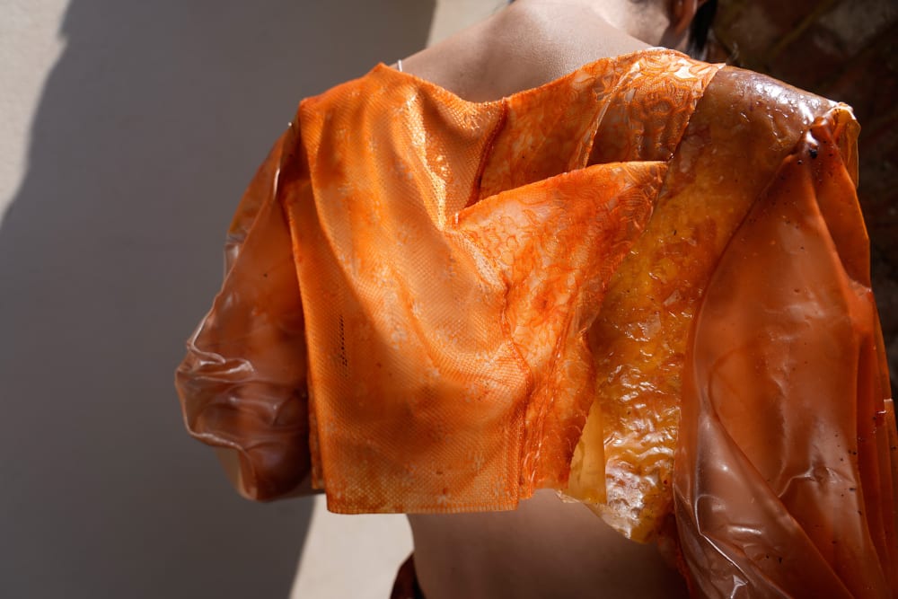A model wears a garment from FATWEAR, the material is textured, involving a lace-like pattern. It is an orange coloured.  