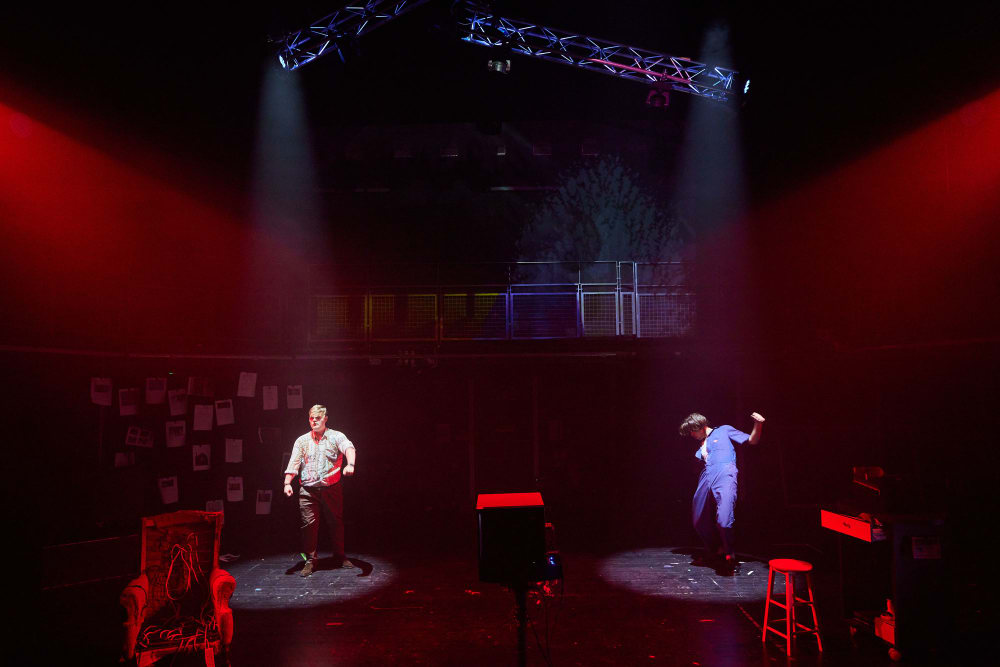 Two men dance on a dark stage. They are each under spotlights and the rest of the stage is lit with red light.
