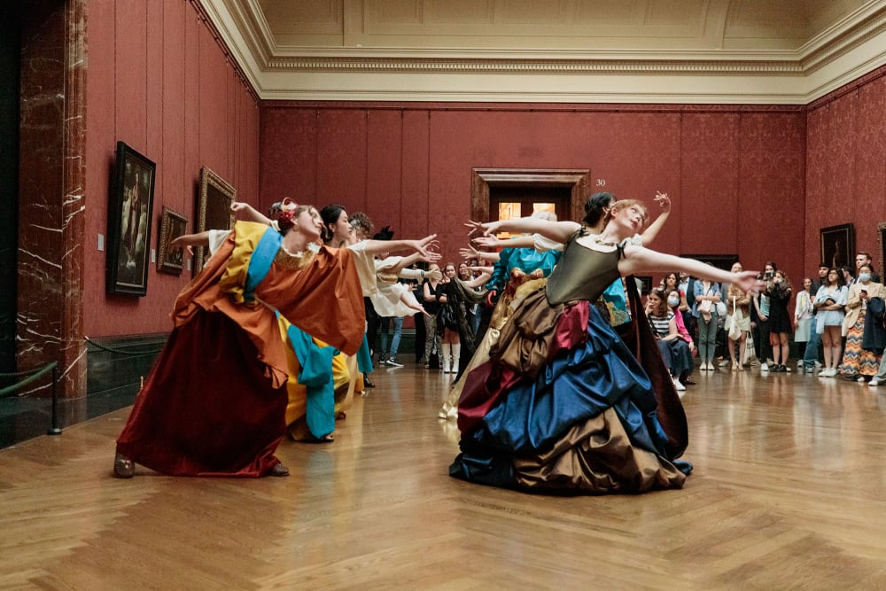 A group of people in costumes do a choreographed performance in a grand gallery space.