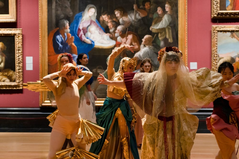 A group of people in costumes perform in front of a large painting.