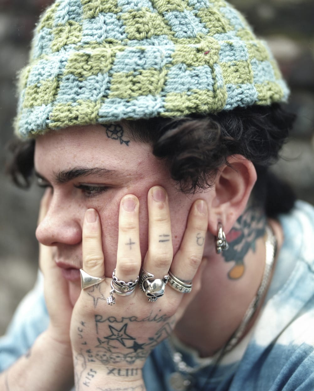 A person rests their face in their hands. Both hands and face are heavily tattooed and adorned in jewellery