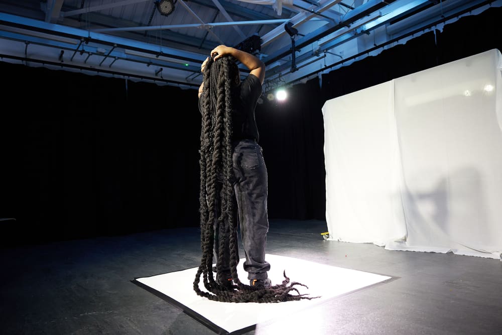 Image shows a young person with long braids standing centre stage with a white screen in the background, slightly to the right. The person is holding their braids as they fall down to the ground.