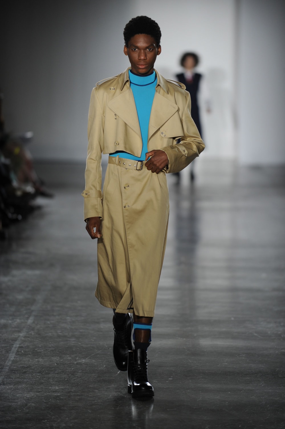 Man on runway wearing cropped beige trench jacket with bright blue turtle neck top and matching trench skirt with belt paired with black boots