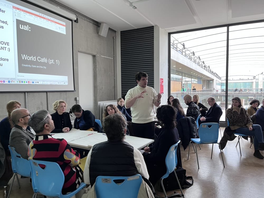 People sitting in groups around tables and one person standing up speaking and presenting thoughts and reflections from the group.