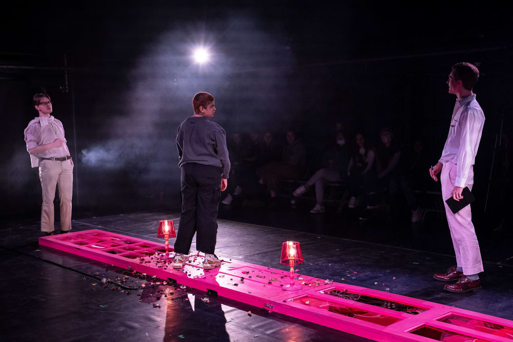 Image shows 3 people standing on stage with 1 person standing on 3 pink doors that are lying horizontally on the ground. The other 2 people stand on either side of the doors.