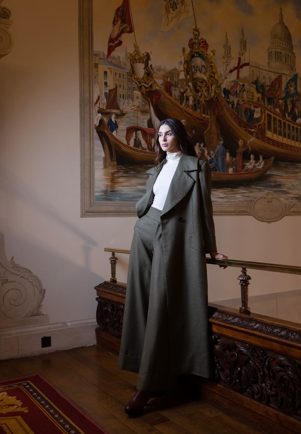 Image shows a person wearing a long dark green jacket and trousers with a white top, posing against a backdrop detailing an ornate painting