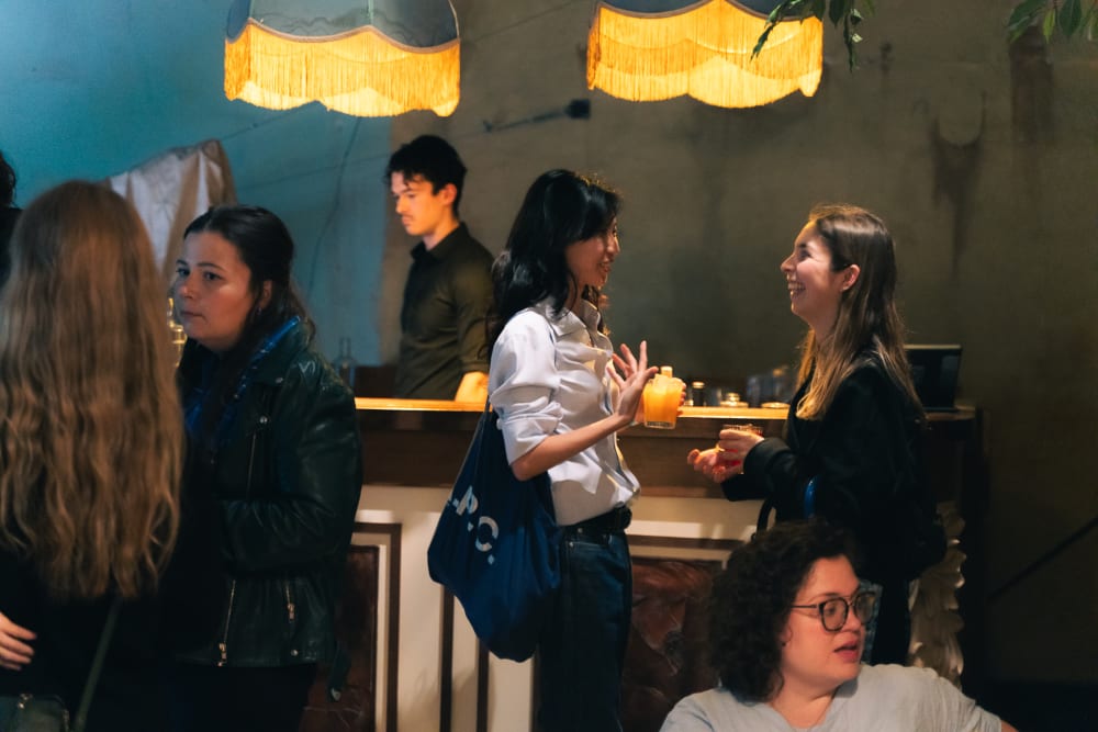 Photo of the room, with people chatting in small groups. 2 yellow lampshades hang down from the ceiling