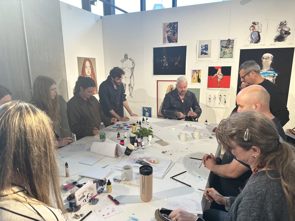 Several people at a table with art equipment on, in a gallery space with artwork on the walls.