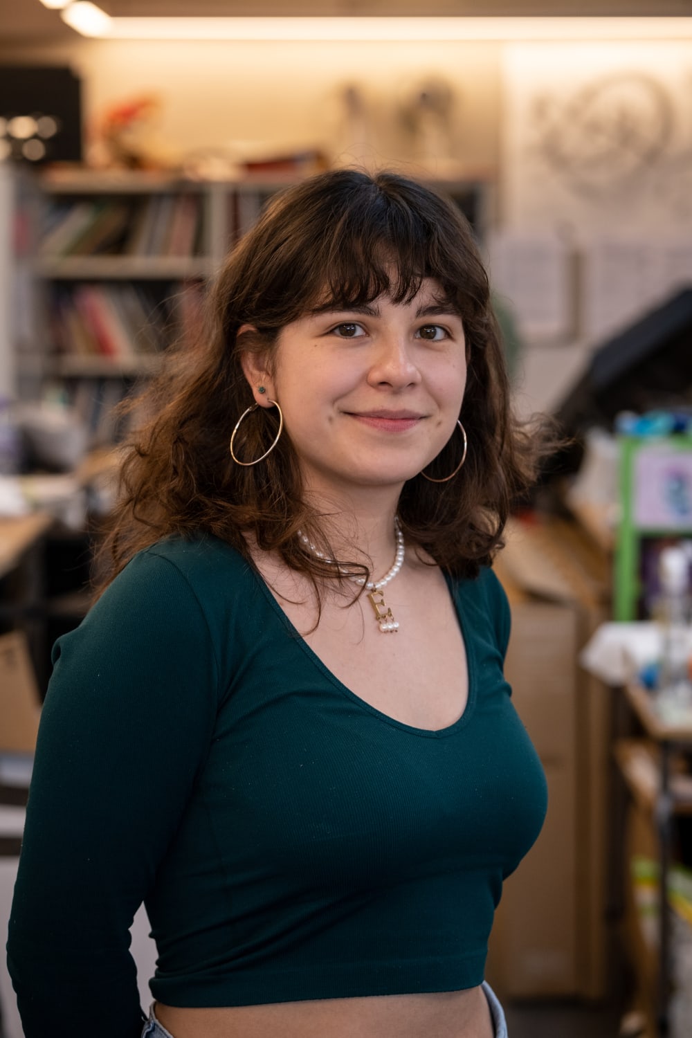 Emilia is smiling into the camera. She has long, brown wavy hair with a fringe. She's wearing a long sleeved top, a pearl necklace with a capital, gold 'E' hanging from it, and large silver hoop earrings.