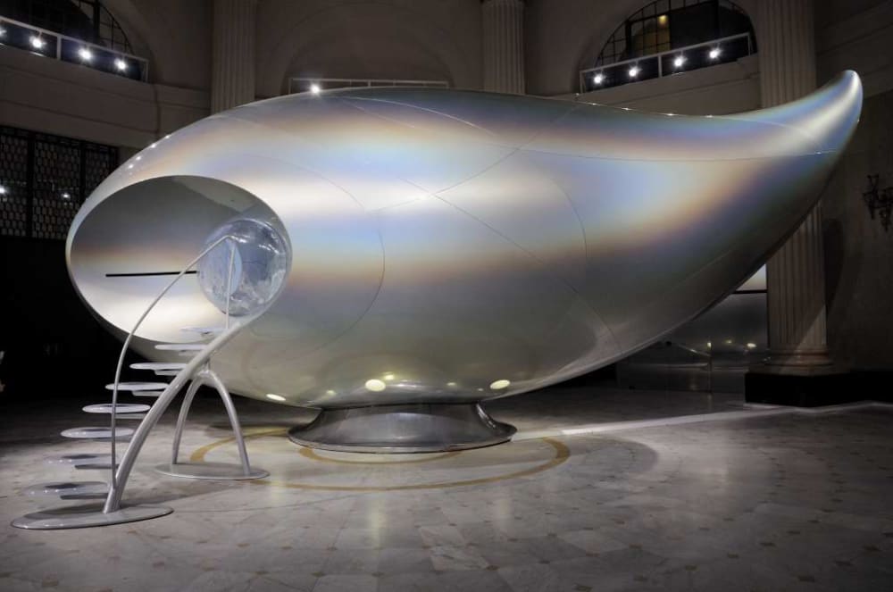 Image shows a large silver structure, mimicking a UFO, with stairs on the left-hand side appearing to lead to an entrance within the structure.