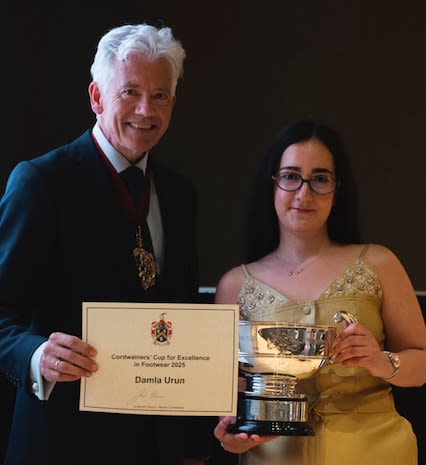 Two people facing the camera to take a picture. One is male with grey hair and a navy blue suit holding a certificate and the other is a female in a gold dress with black hair and glasses holding a trophy. 