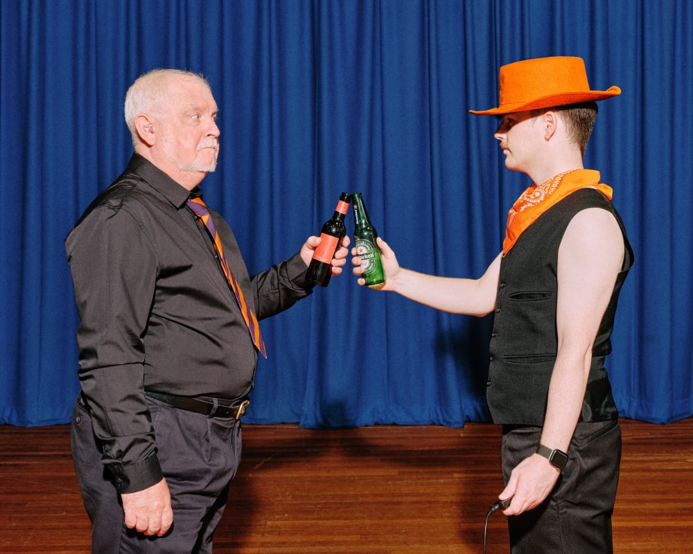 Neil and Ciaran stand on a stage, facing each other. They hold out bottles of beer in a toast