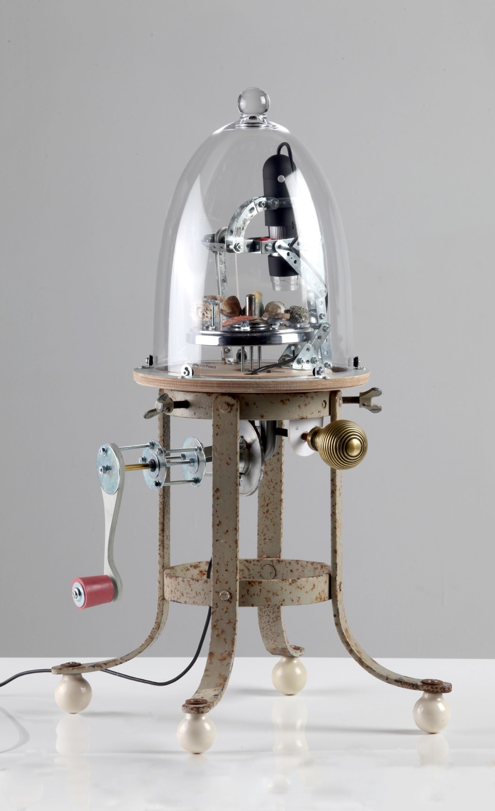 A mechanical contraption under a glass dome, sits on a set of four metal legs which are raised off a table surface by pearls.