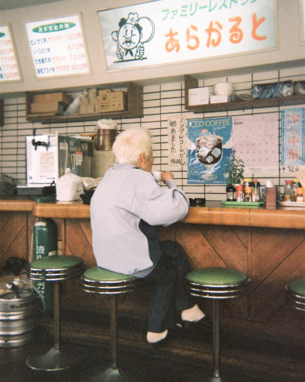There is a man, with his back to us sitting on a stool at a bar drinking from a white cup. The wall behind the bar is decorated with advertisements and you can see condiments neatly lined up. 