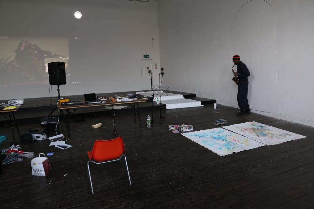 A room with a projection of a video showing on the white wall. A male saxophonist is playing in the corner.