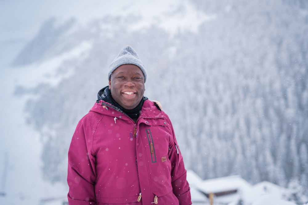 A man in a blue wooly hat and a purple warm jacket stands smiling at the camera in a snowy, mountainous landscape.