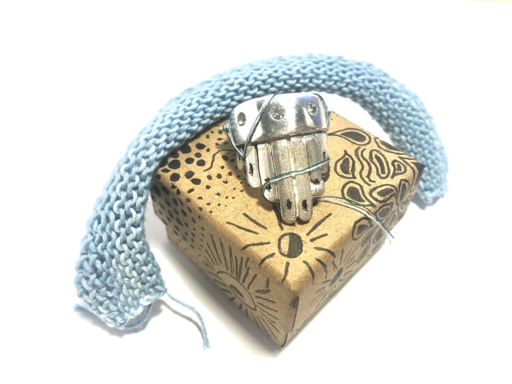 A silver ring shaped like a hand is positioned on a decorative ring box, with a piece of blue knitted material draped over the piece.  