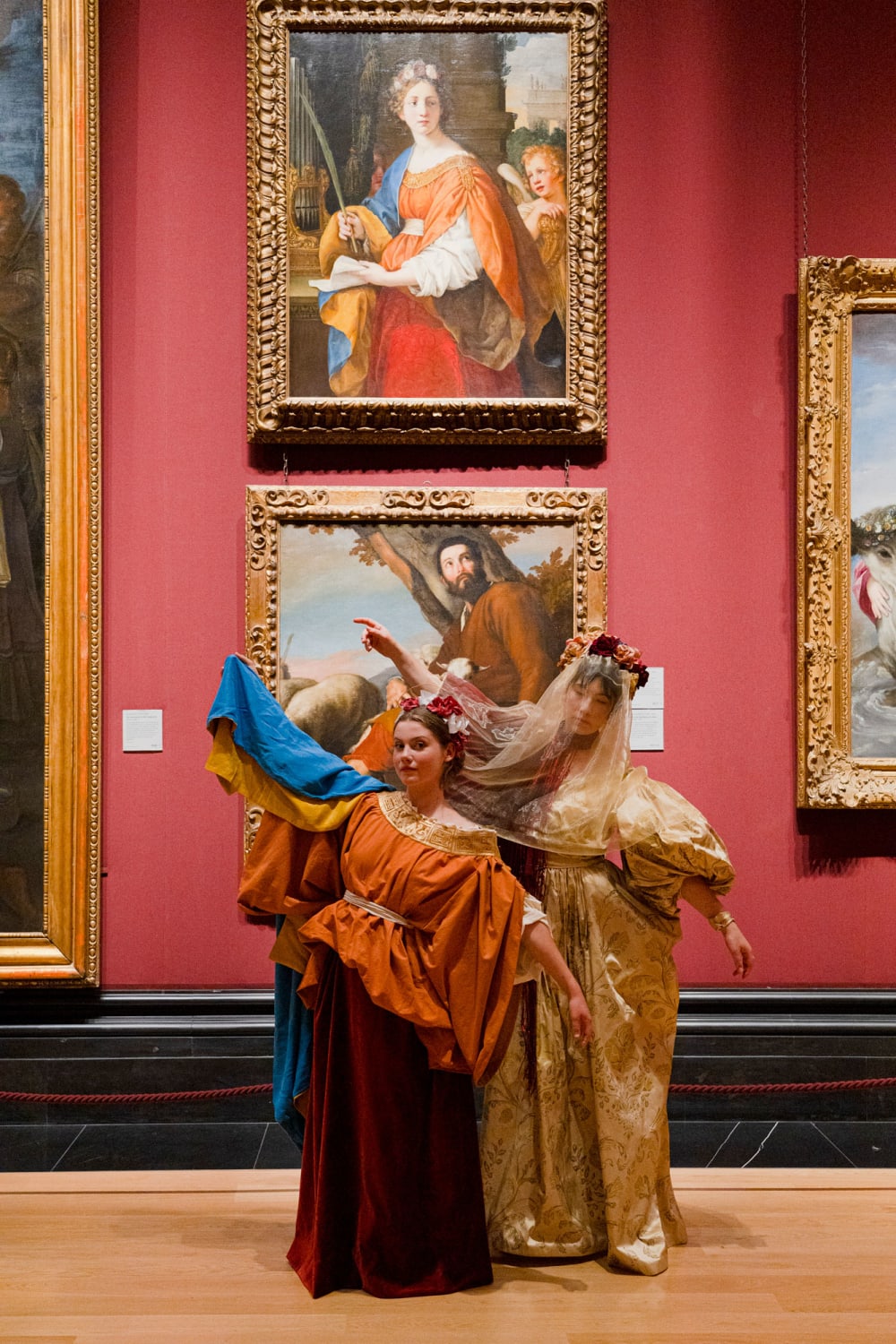 Two young women in costumes stand in front of two paintings in gold frames hanging on a red wall.