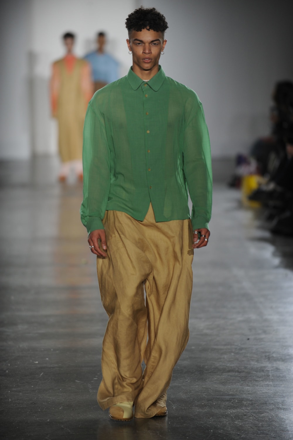 Man on runway wearing chiffon sage green shirt and baggy flowy beige trousers and silver jewellery: hooped earrings, chocker and rings
