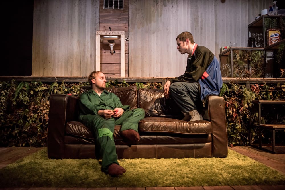 Two men sitting on a brown leather sofa; one is sat on the right arm with his feet on the seat, and the other is sat on the far left of the sofa. They're looking at each other, talking. Behind them a stage is covered in green foliage, and there's a green rug on the floor.