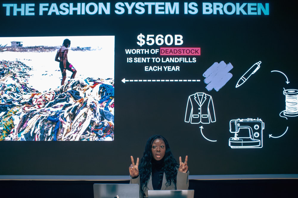 Close up photo of Giovanna presenting. On the screen behind her it says: the fashion system is broken $560B worth of deadstock is sent to landfills each year. 
