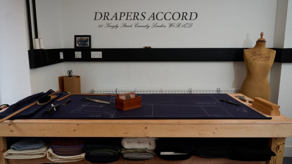 A pattern cutting table with a dress makers mannequin to the right, a pair of scissors on top in front of a white wall with Drapers Accord written in vinyl