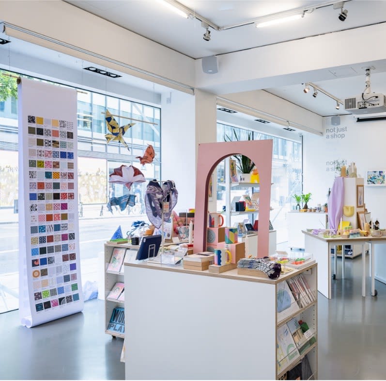 Interior view of the 'not just a shop' retail space at UAL. The photo shows a bright, modern shop with white walls, large windows, and product displays. In the left foreground is a large vertical banner featuring a grid of colourful printed patterns, placed in front of the shop’s window display area facing the street. Hanging paper sculptures resembling origami are suspended in the window above. Display tables and shelving units hold a range of colourful products, including mugs, books, prints, and stationery. A pink arch-shaped mirror is positioned on one of the central units, and the back wall features the 'not just a shop' logo.