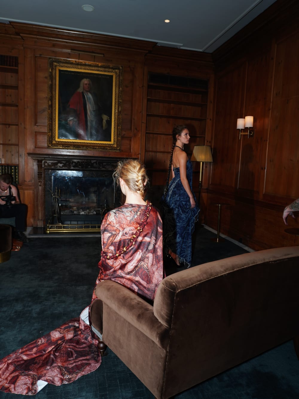 Two models in elegant gowns pose in a wood-paneled room with a portrait above a fireplace. One sits on a couch; the other stands, creating a serene atmosphere.