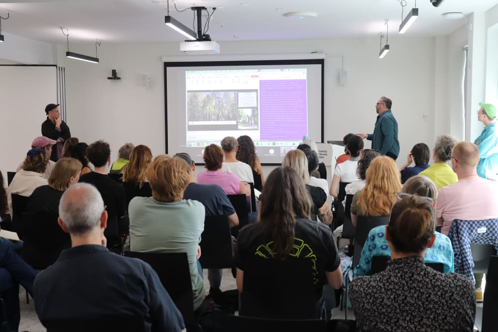 Dylan Robinson by projected slides at lectern reading from paper. Backs of audience heads.