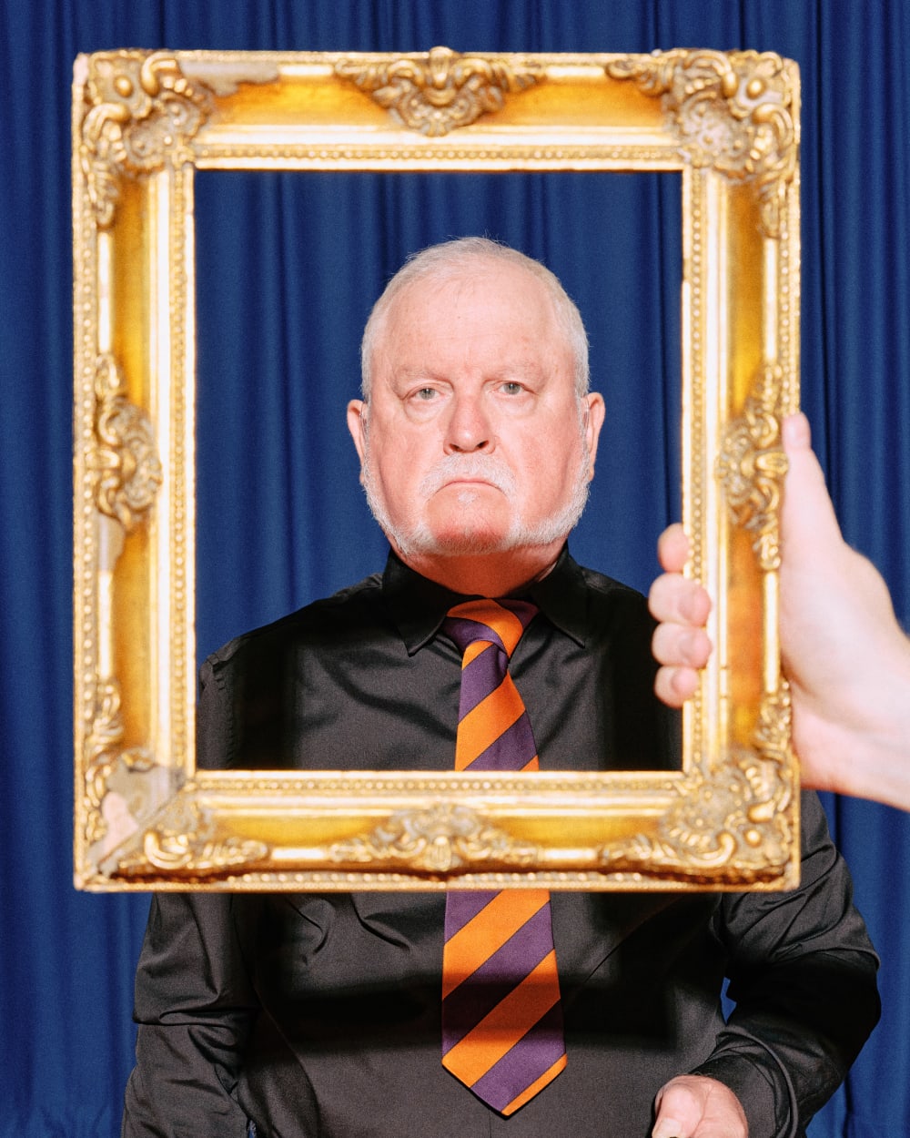 A hand holds a golden frame in front of Neil, who stares neutrally into the camera