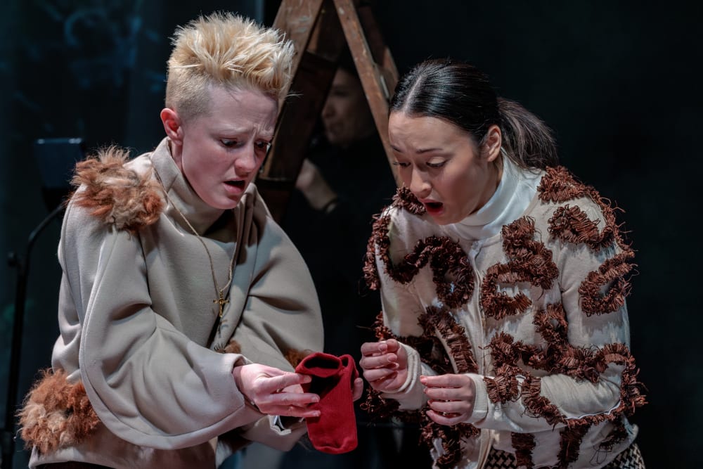 Two individuals in textured, costume-style garments stand closely together on stage while holding and looking at a small red fabric object.