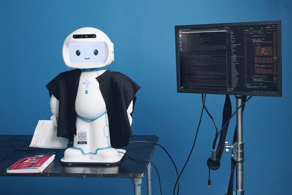 A small robot in graduation robes sits on a table next to a screen with code on it.
