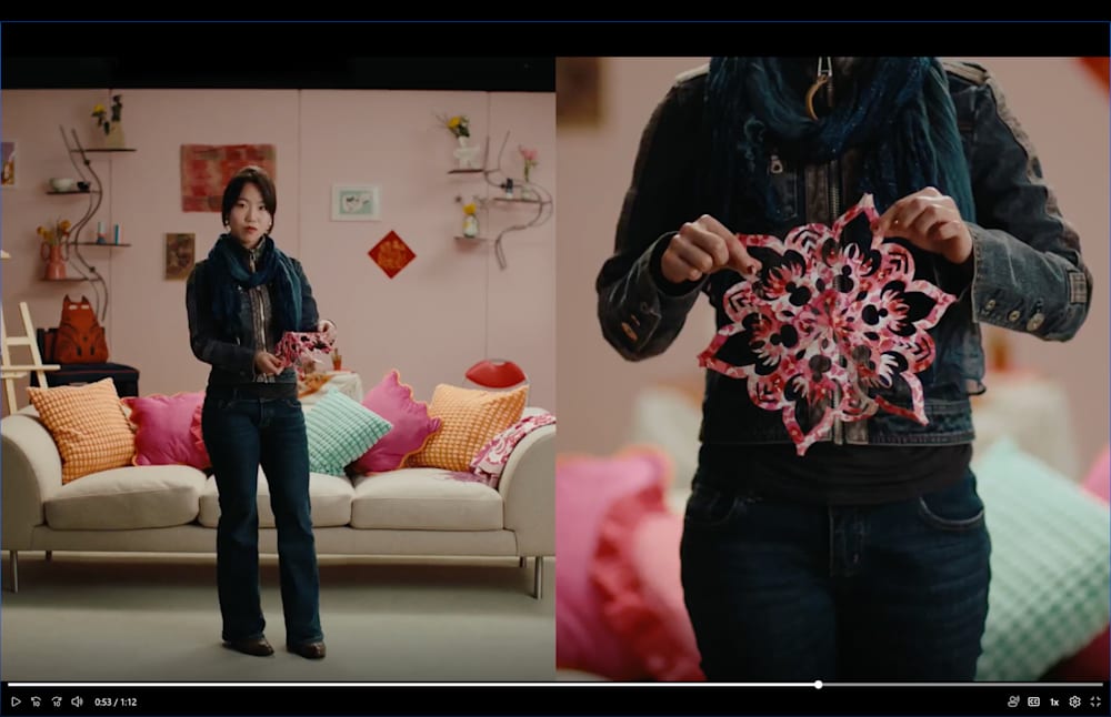 A split-screen video still showing a young woman standing in a colourful studio set styled as a living room, holding a decorative paper-cut artwork. The right panel shows a close-up of the same piece — an intricate floral design in red, pink, and black.