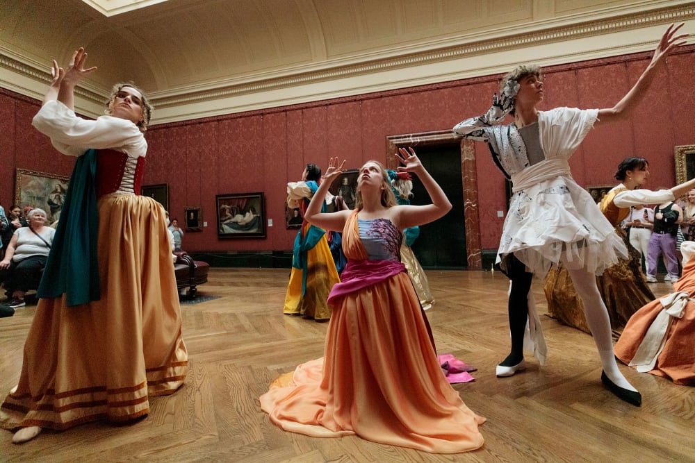 Performers in different poses face in different dircetions in a large gallery space with paintings behind them.