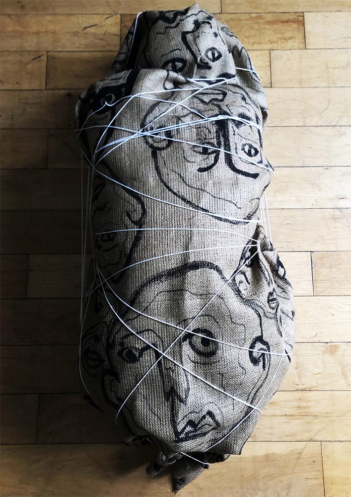 a person wrapped in a hessian sack covered in string and drawings of faces 