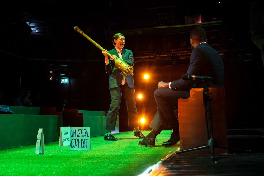 Image shows 2 people in a set designed to look like the United Kingdom's House of Commons. It is green with benches on either side. 1 person is holding a large mace and swinging it towards another person who can be seen sitting down on the right hand side bench. Small placards can be seen on the ground reading 'Tax Loopholes' and 'Universal Credit'.