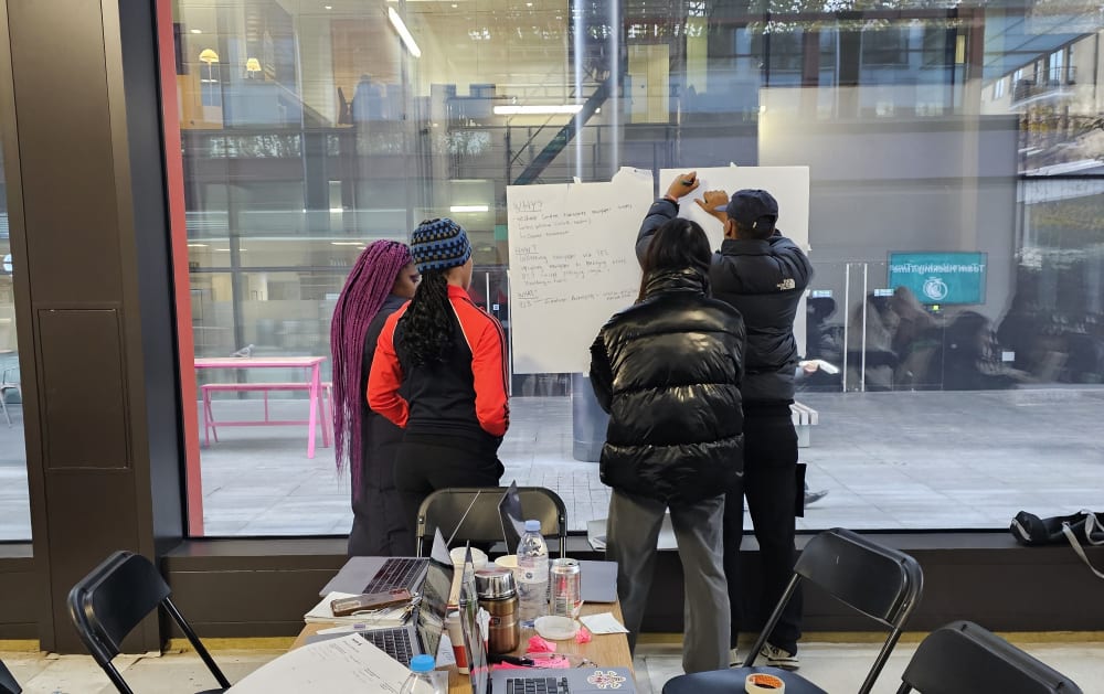 $ students standing around 2 pieces of A3 paper, taped to a window. 
