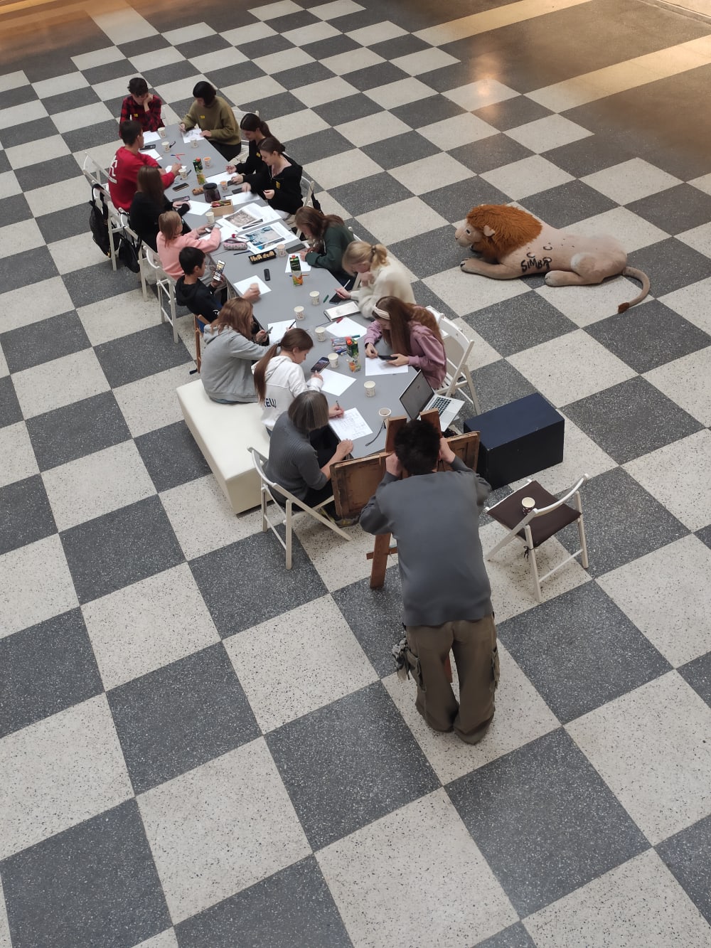 A shot of the participants at work at a long table, shot sideways and from above. The floor is check tiled grey and white and a stuffed lion is visible to the right