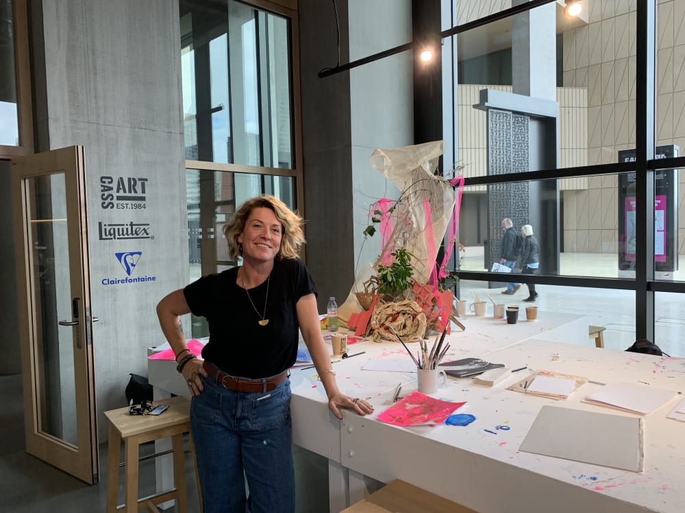Person smiling at camera, leaning on table with art supplies on,