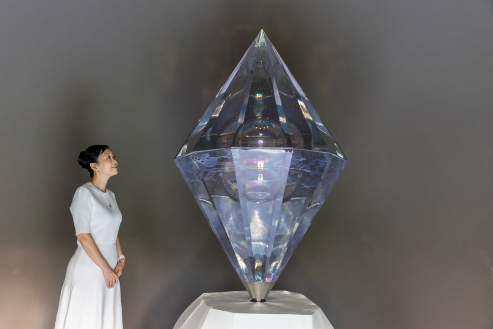 Image shows a woman dressed in white standing to the left of a large installation of a “diamond” crystal. The woman is smiling while looking up at the diamond.