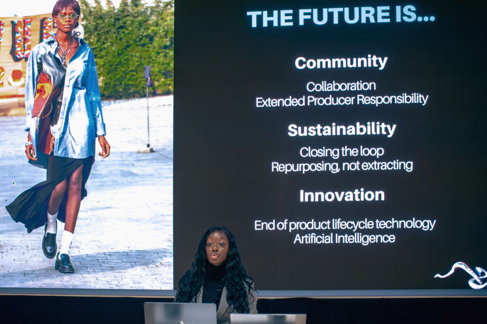 Close up photo of Giovanna speaking. On the screen behind her is a slide with a photo of model walking on the left. On the right it says: THE FUTURE IS... Community, collaboration, extended producer responsibility; Sustainability, closing the loop, repurposing, not extracting; Innovation, end of product lifecycle technology, Artificial Intelligence