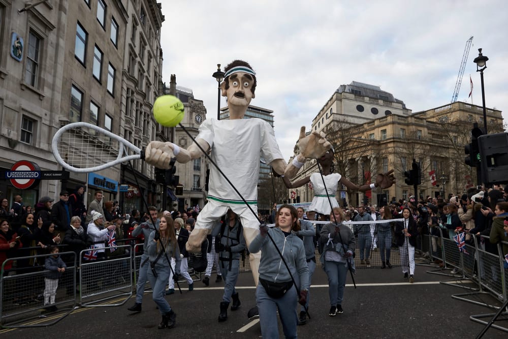 Two large puppets of tennis players are operated by people in grey tracksuits in front of large crowds in central London.