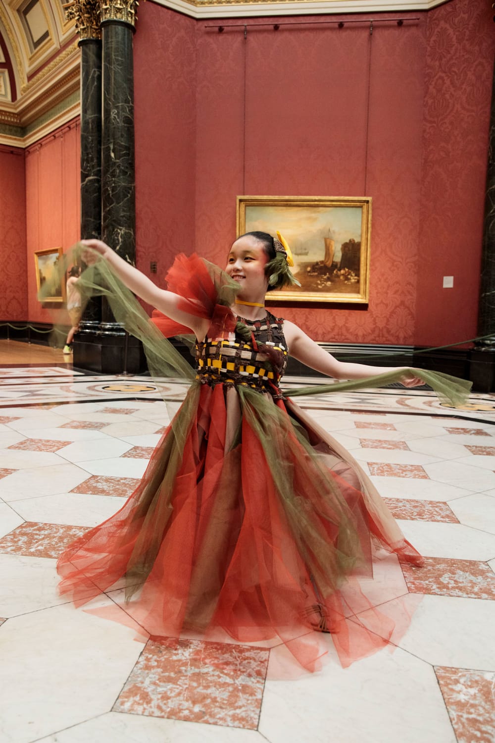 A woman stands in a large gallery space with a marble floor. A painting is hanging on a red wall behind her. She is wearing a red and green net dress with a plastic bodice. Her arms are outstretched.