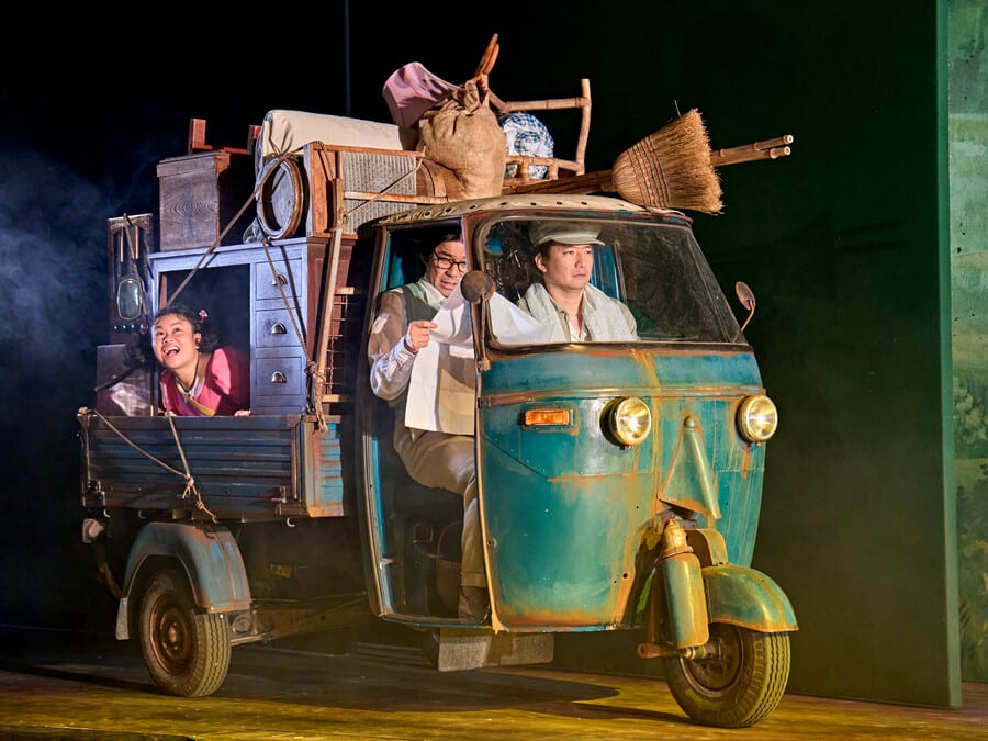 Image shows a tuk-tuk with household items on top. There are 2 people driving and an actor playing a young child in the back of the vehicle with her head sticking out of the window